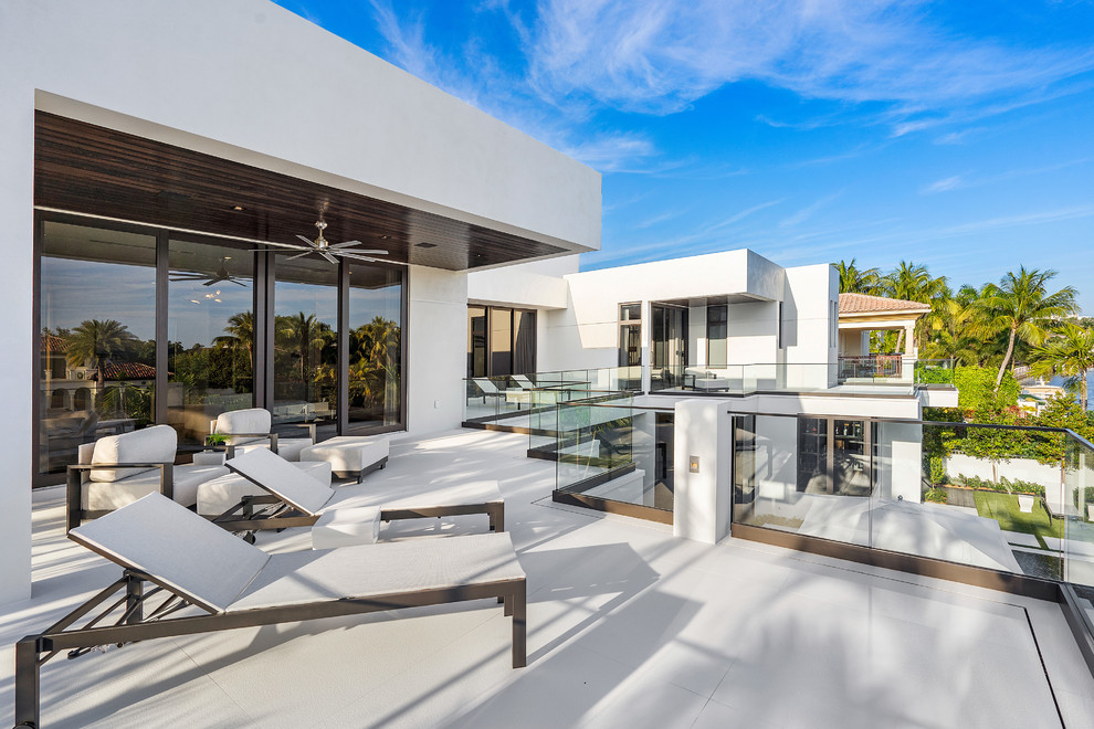 تراس مدرن ویلا با طراحی سفید، مبلمان فضای باز، نرده شیشه‌ای و چشم‌انداز سبز در معماری معاصر مناطق گرمسیری.

