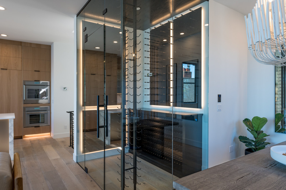 اتاق شیشه‌ای مدرن برای نگهداری نوشیدنی با قفسه‌های مشکی، نورپردازی داخلی و طراحی لوکس در کنار آشپزخانه چوبی
