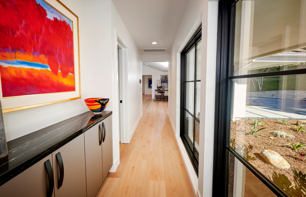 راهروی مدرن با کفپوش چوبی روشن، پنجره‌های بزرگ مشکی، تابلو هنری رنگارنگ و کابینت سنگی مشکی برای دکور داخلی خانه

