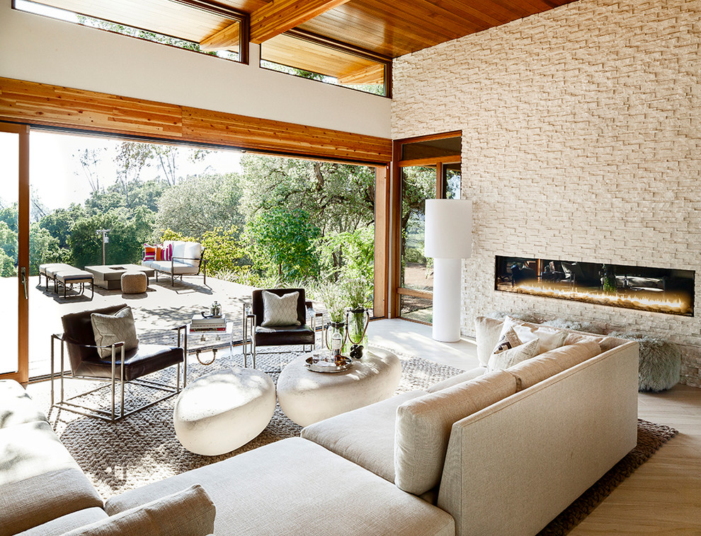 طراحی نشیمن مدرن و ارگانیک با دیوار سنگی سفید، شومینه خطی، مبل کرم رنگ، میزهای عسلی سنگی، نور طبیعی فراوان و دسترسی به تراس فضای باز