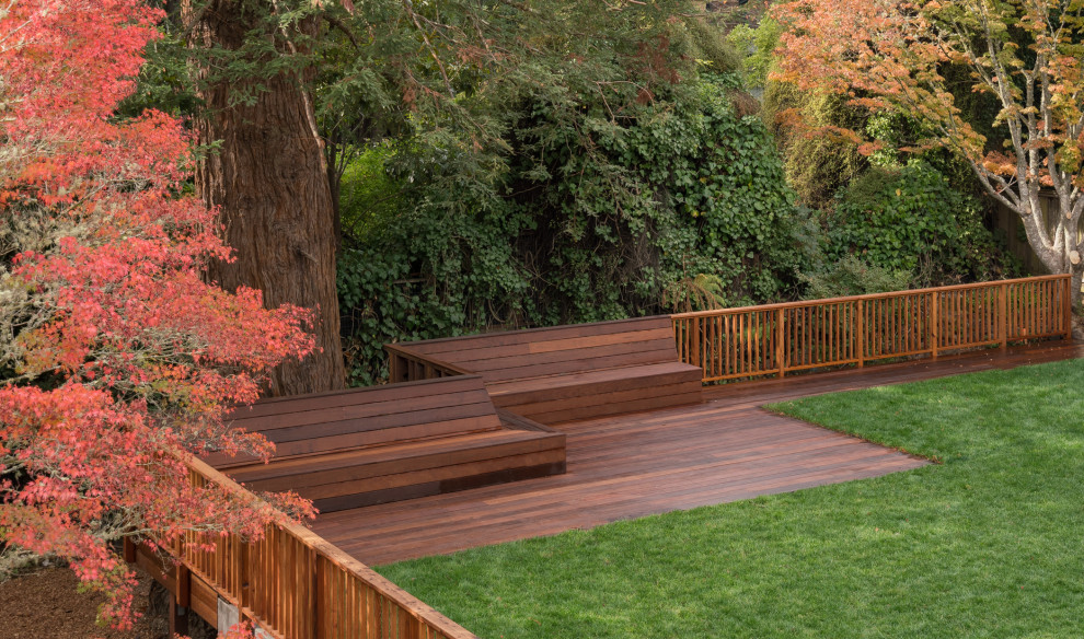 طراحی کلاسیک حیاط پشتی بزرگ با فضای چوبی وسیع، نیمکت‌های یکپارچه چوبی، نرده‌های چوبی ساده، چمن سبز و درختان پاییزی با برگ‌های رنگارنگ.