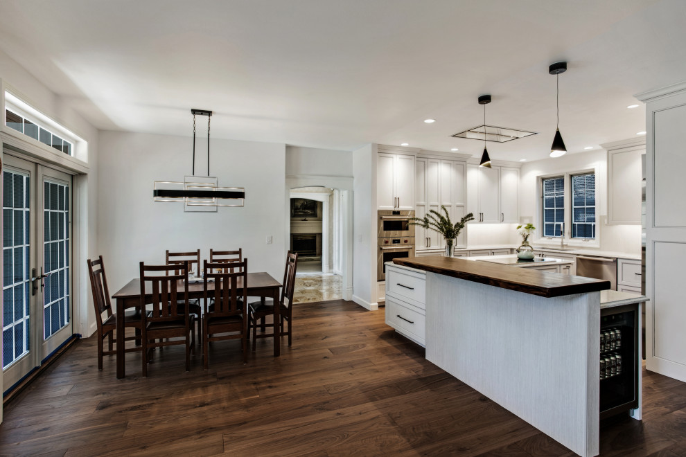 طراحی داخلی آشپزخانه مدرن با جزیره چوبی، کابینت‌های سفید، نورپردازی معلق و فضای ناهارخوری با میز و صندلی چوبی کلاسیک