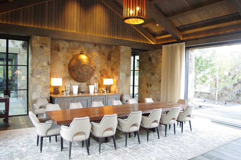 سالن ناهارخوری با سقف چوبی شیب‌دار، دیوار سنگی، میز بزرگ چوبی و نور طبیعی در طراحی داخلی لوکس به سبک روستیک مدرن