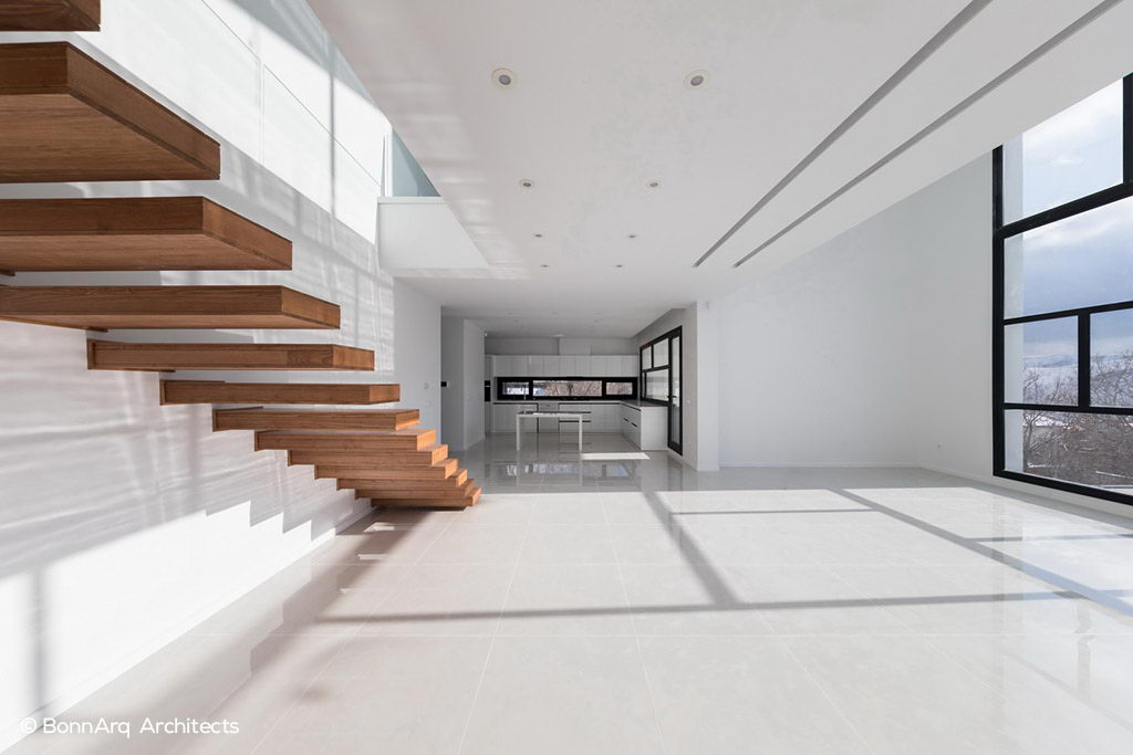 نمای داخلی خانه مدرن با پلکان معلق چوبی، آشپزخانه یکپارچه سفید، پنجره‌ بلند و نور طبیعی گسترده بر کف براق.