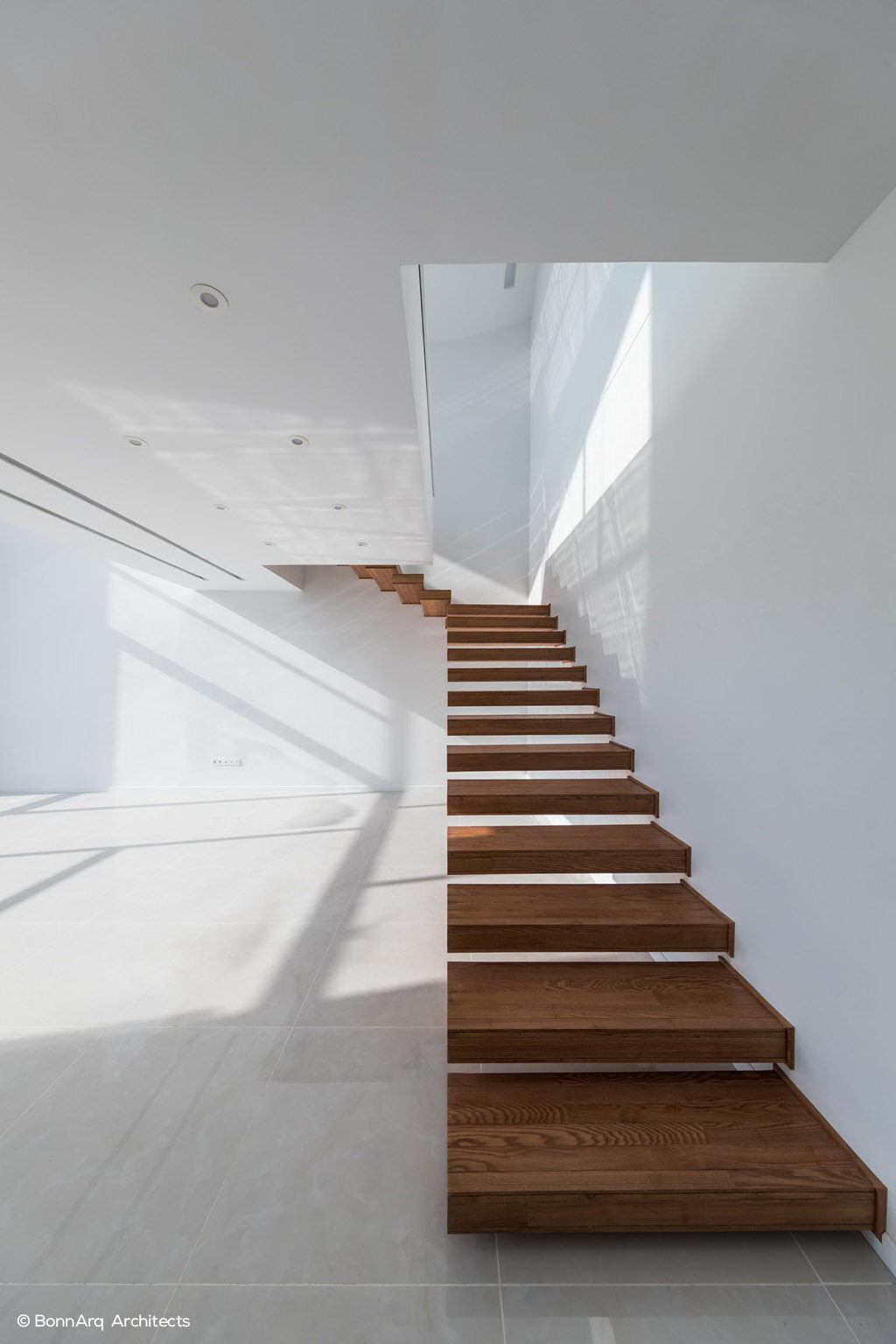 نمای نزدیک پلکان معلق چوبی در فضای داخلی سفید با نور طبیعی گسترده، طراحی مدرن، مینیمال و بازی سایه در معماری.