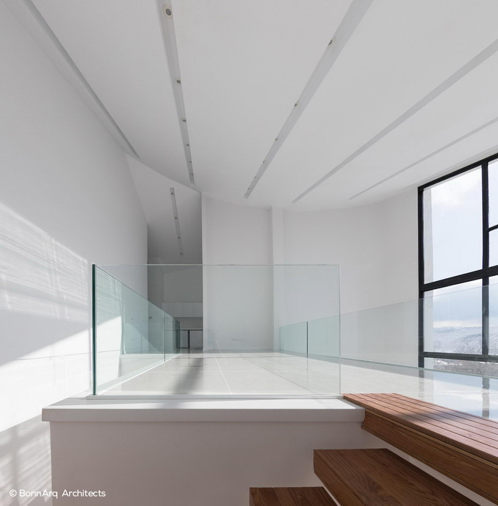 نمای طبقه دوم خانه مدرن با نرده شیشه‌ای، نور طبیعی گسترده، سقف منحنی و پله‌های چوبی در فضای روشن و مینیمال.