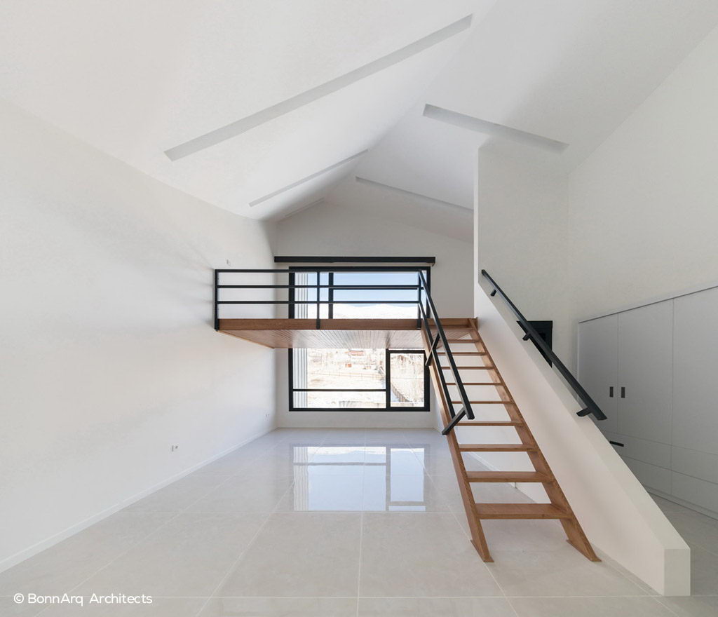 اتاق‌خواب طبقه دوم با سقف شیبدار، نیم‌طبقه چوبی با نرده فلزی، نور طبیعی از پنجره بزرگ و طراحی مدرن و ساده.