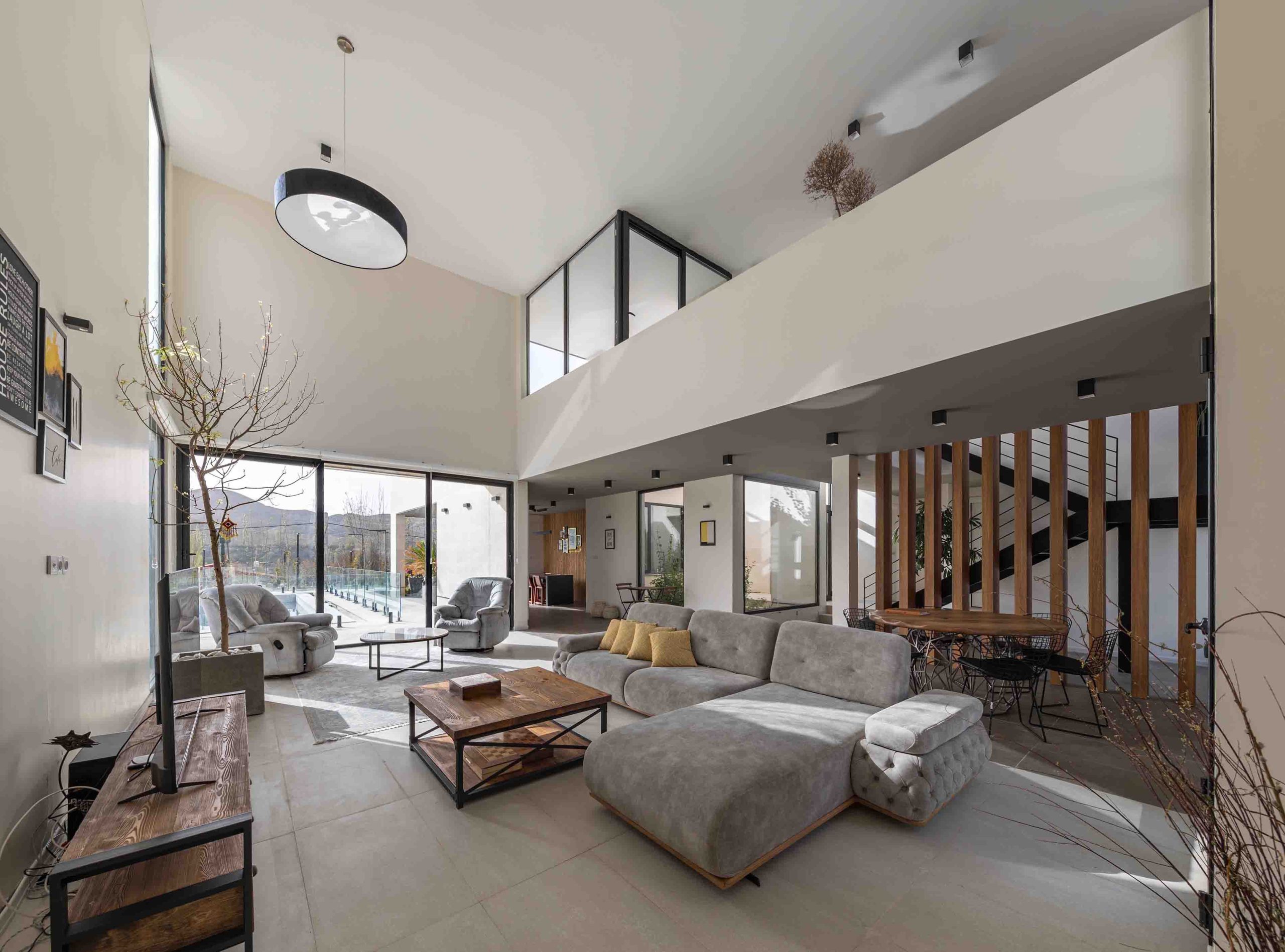 پلان باز ویلای دوبلکس با نور طبیعی، مبلمان طوسی، پارتیشن چوبی و پنجره‌های قدی مشرف به استخر و فضای بیرونی