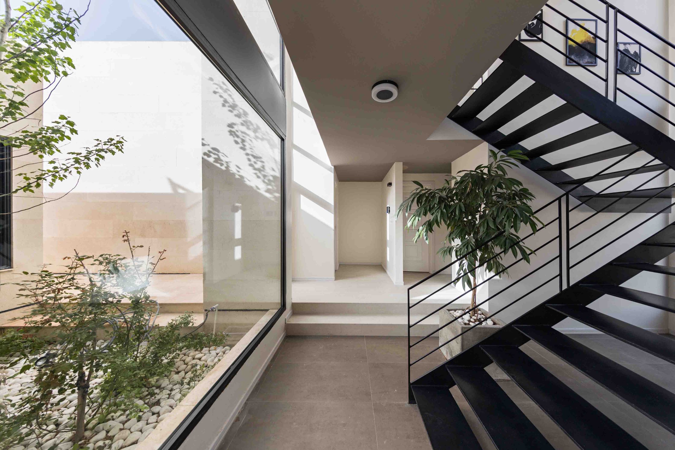 طراحی مینیمال فضای راه‌پله با نرده فلزی مشکی، گیاهان طبیعی، نورگیر بزرگ و ترکیب سنگ، سبز و نور در معماری داخلی